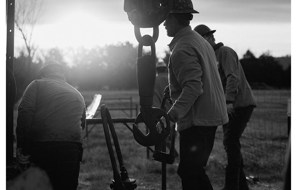Varios trabajadores con cascos y chaquetas operan maquinaria pesada al aire libre al amanecer o al atardecer. Un trabajador maneja un gancho industrial grande, mientras otros ayudan en una zona cercada con césped.